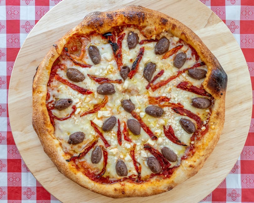 A whole pizza with olives, sun-dried tomatoes, and feta cheese on a wooden board.