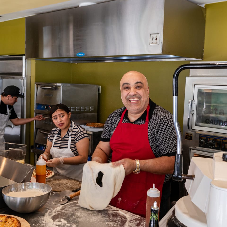 Smiling chef in a red apron stretching pizza dough, with two other chefs working in the background.