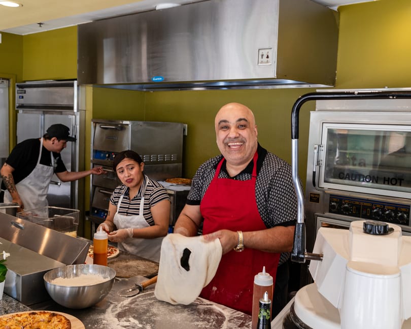 Smiling chef in a red apron stretching pizza dough, with two other chefs working in the background.