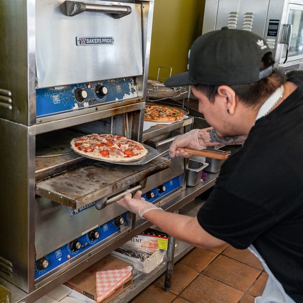 Chef in black cap and gloves placing a pepperoni and bacon pizza into a Bakers Pride oven.