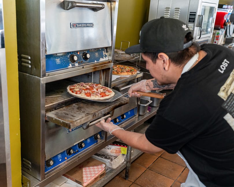 Chef in black cap and gloves placing a pepperoni and bacon pizza into a Bakers Pride oven.