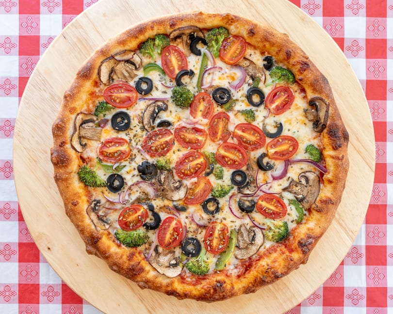 A whole vegetarian pizza with tomatoes, olives, mushrooms, broccoli, and onions on a wooden board.