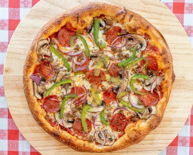 A whole pizza with pepperoni, sausage, mushrooms, green peppers, and red onions on a wooden board.