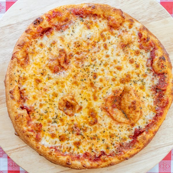 A whole cheese pizza with herbs on a wooden board over a red and white checkered tablecloth.