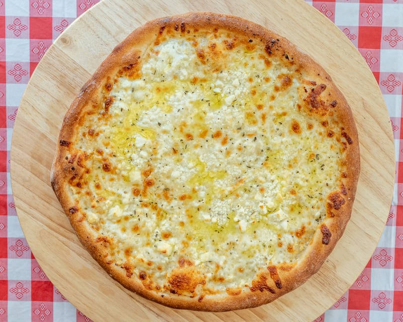 White pizza with ricotta, mozzarella, olive oil, and herbs on a wooden peel over a red and white tablecloth.