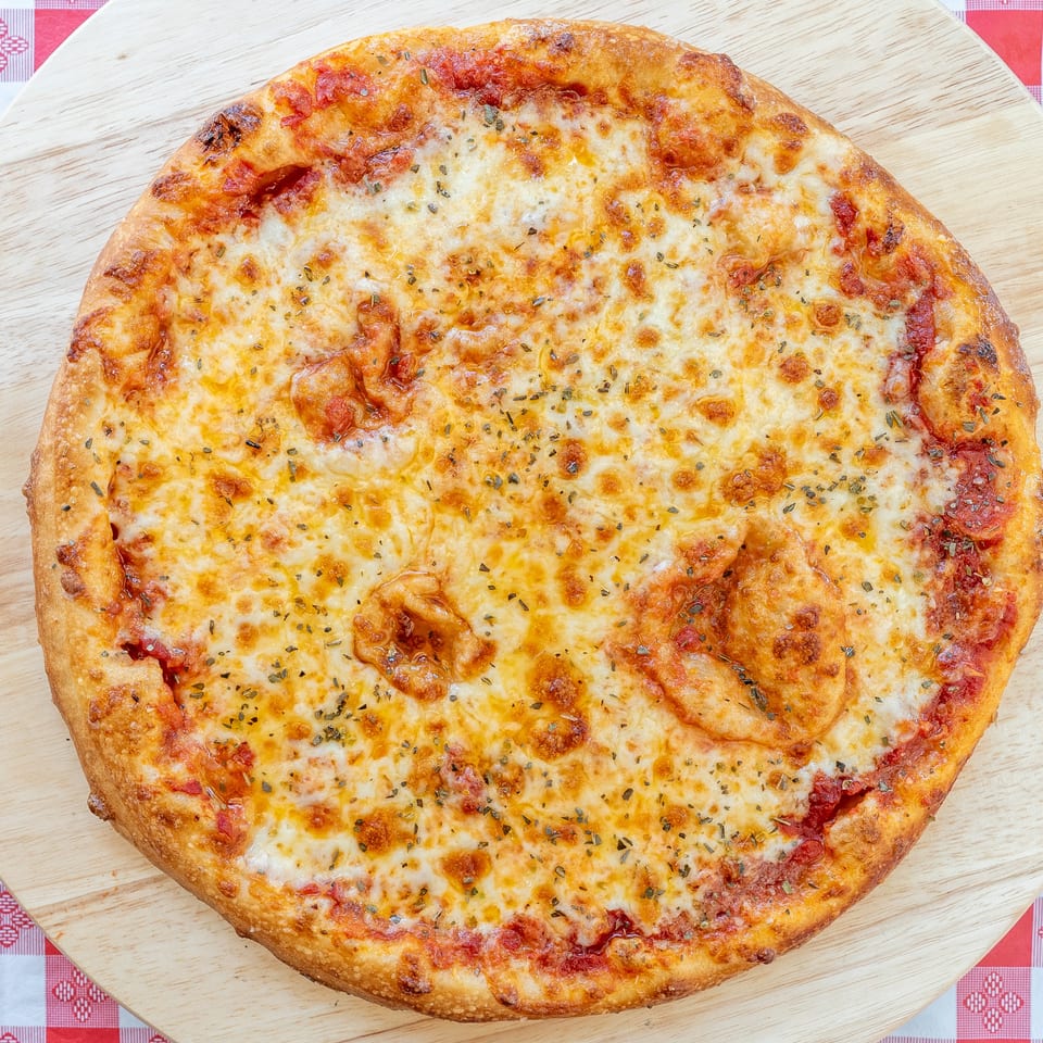 A whole cheese pizza with herbs on a wooden board over a red and white checkered tablecloth.