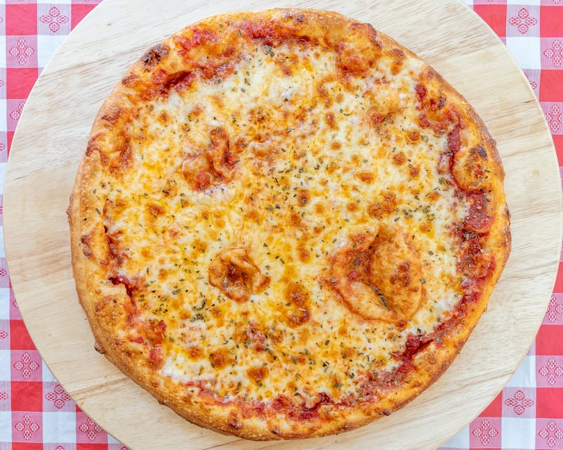 A whole cheese pizza with herbs on a wooden board over a red and white checkered tablecloth.