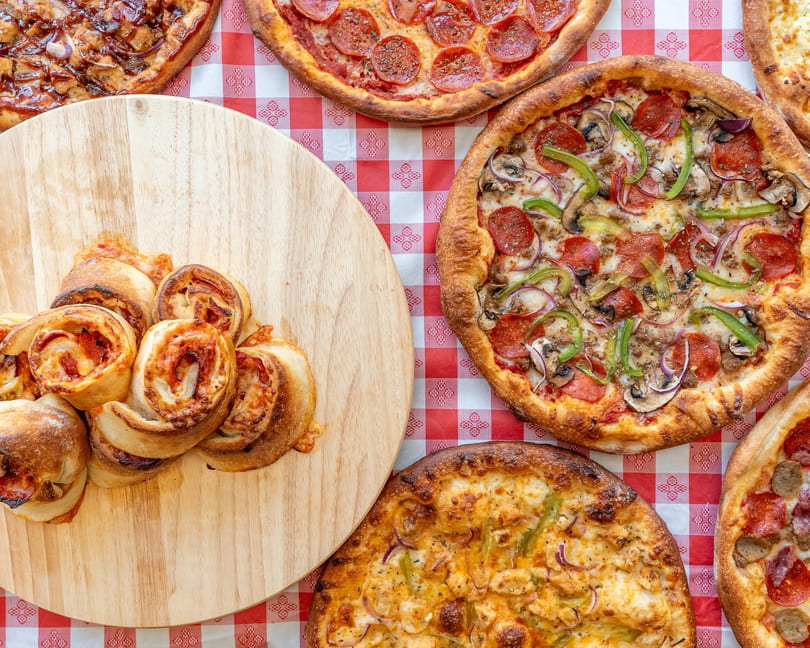 Assortment of pizzas and pepperoni rolls on a red and white checkered tablecloth.
