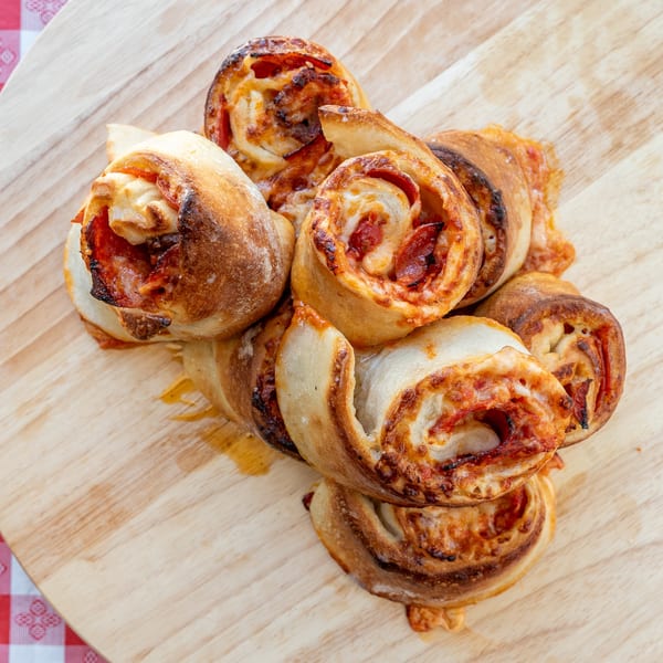 Golden-brown pizza rolls with pepperoni and cheese, piled on a light wooden board next to a red and white checkered cloth.