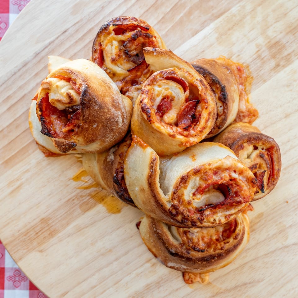 Golden-brown pizza rolls with pepperoni and cheese, piled on a light wooden board next to a red and white checkered cloth.