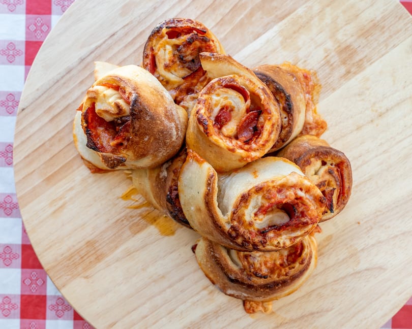 Golden-brown pizza rolls with pepperoni and cheese, piled on a light wooden board next to a red and white checkered cloth.
