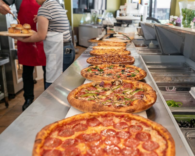 A row of freshly baked pizzas with various toppings on a stainless steel counter in a restaurant kitchen.