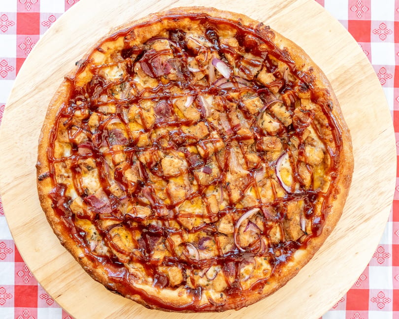 BBQ chicken pizza with red onions and bacon, drizzled with sauce, on a wooden board.