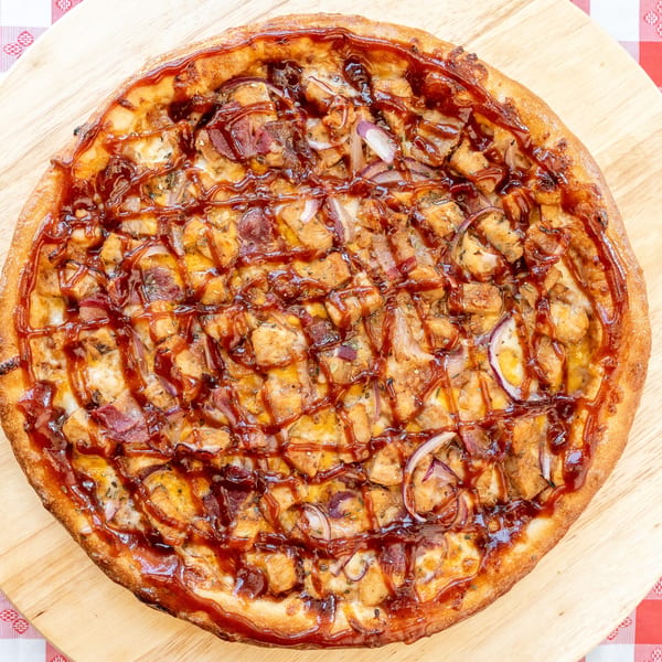 BBQ chicken pizza with red onions and bacon on a wooden board over a red and white checkered tablecloth.
