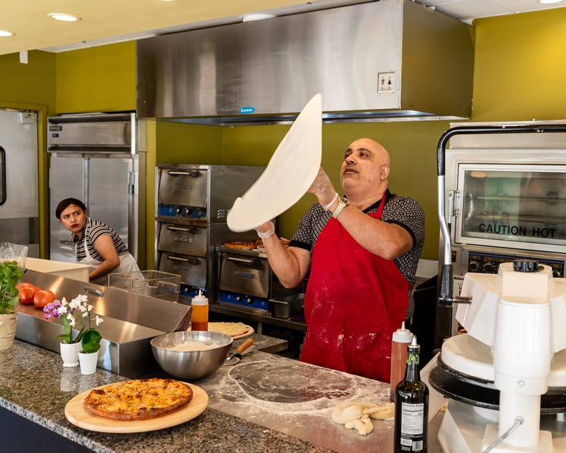 A chef in a red apron expertly tosses pizza dough in a bustling kitchen with another worker in the background.