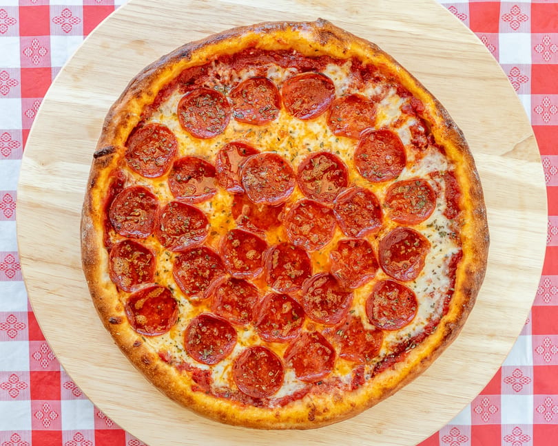 A whole pepperoni pizza with a thick crust on a wooden board and red checkered tablecloth.