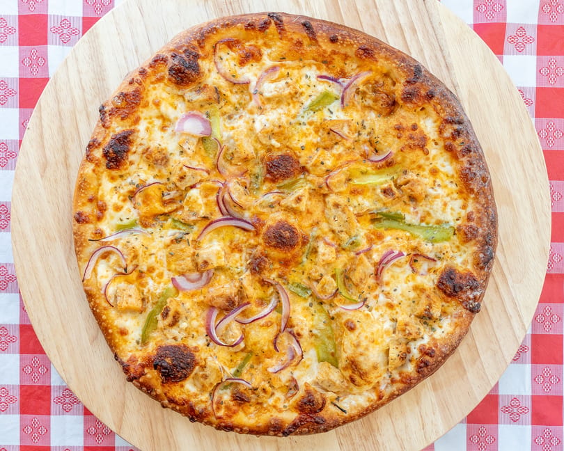 Overhead view of a whole chicken and vegetable pizza on a wooden board, on a red and white checkered tablecloth.
