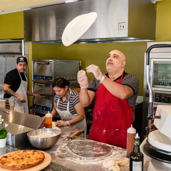 Chef in red apron tossing pizza dough in the air, with other chefs working in the background.