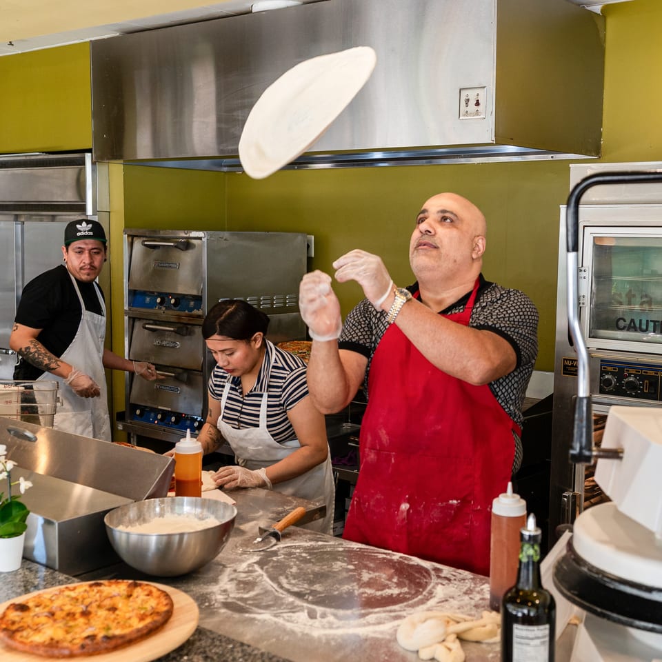 Chef in red apron tossing pizza dough in the air, with other chefs working in the background.