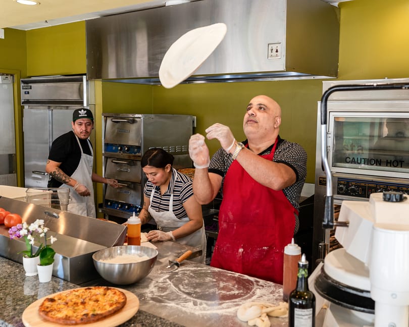 Chef in red apron tossing pizza dough in the air, with other chefs working in the background.