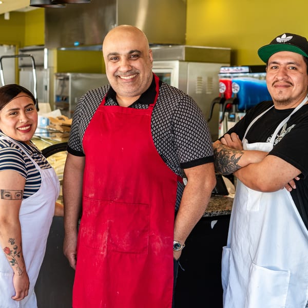 Three smiling restaurant staff members, two men and one woman, wearing aprons in a kitchen.