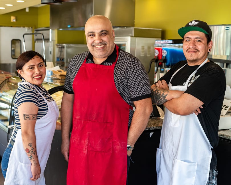 Three smiling restaurant staff members, two men and one woman, wearing aprons in a kitchen.