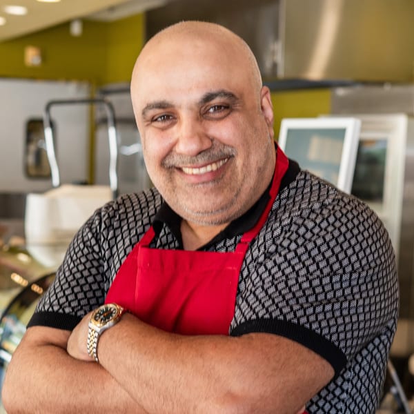 Smiling bald man with a mustache wearing a red apron and patterned shirt, arms crossed.