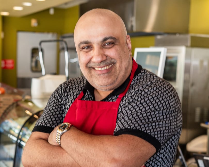 Smiling bald man with a mustache wearing a red apron and patterned shirt, arms crossed.
