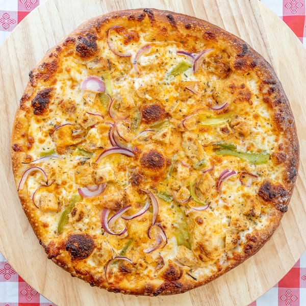 A whole chicken, onion, and green pepper pizza on a wooden peel over a red and white checkered tablecloth.
