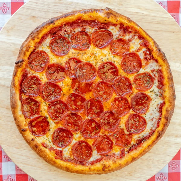 A whole pepperoni pizza with a thick crust on a wooden board and red checkered tablecloth.