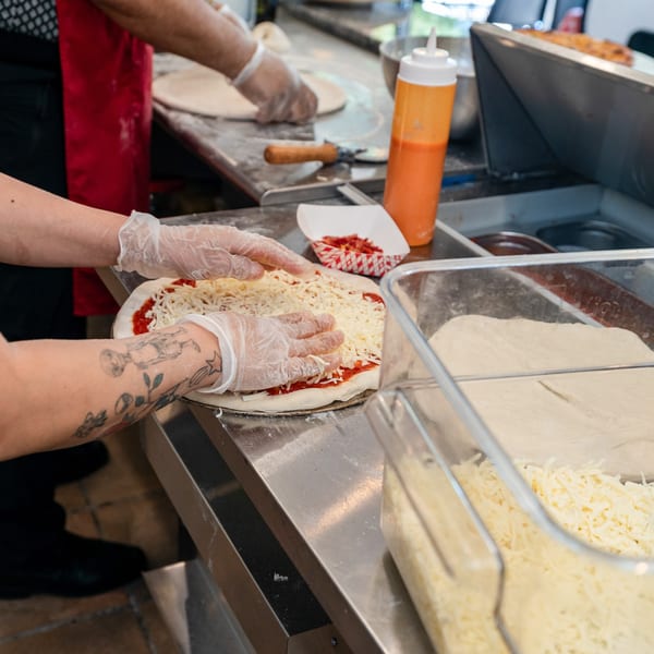 Gloved hands spread shredded cheese on pizza dough with sauce, next to containers of ingredients.