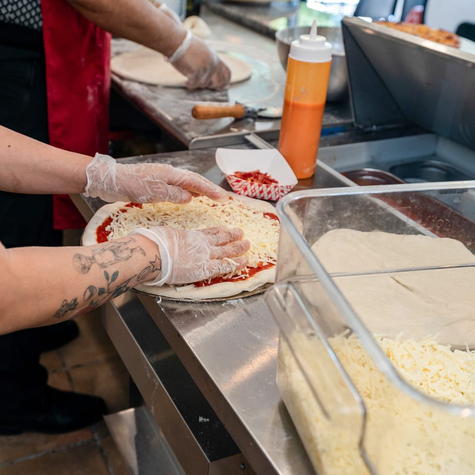 Gloved hands spread shredded cheese on pizza dough with sauce, next to containers of ingredients.