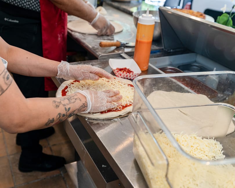 Gloved hands spread shredded cheese on pizza dough with sauce, next to containers of ingredients.